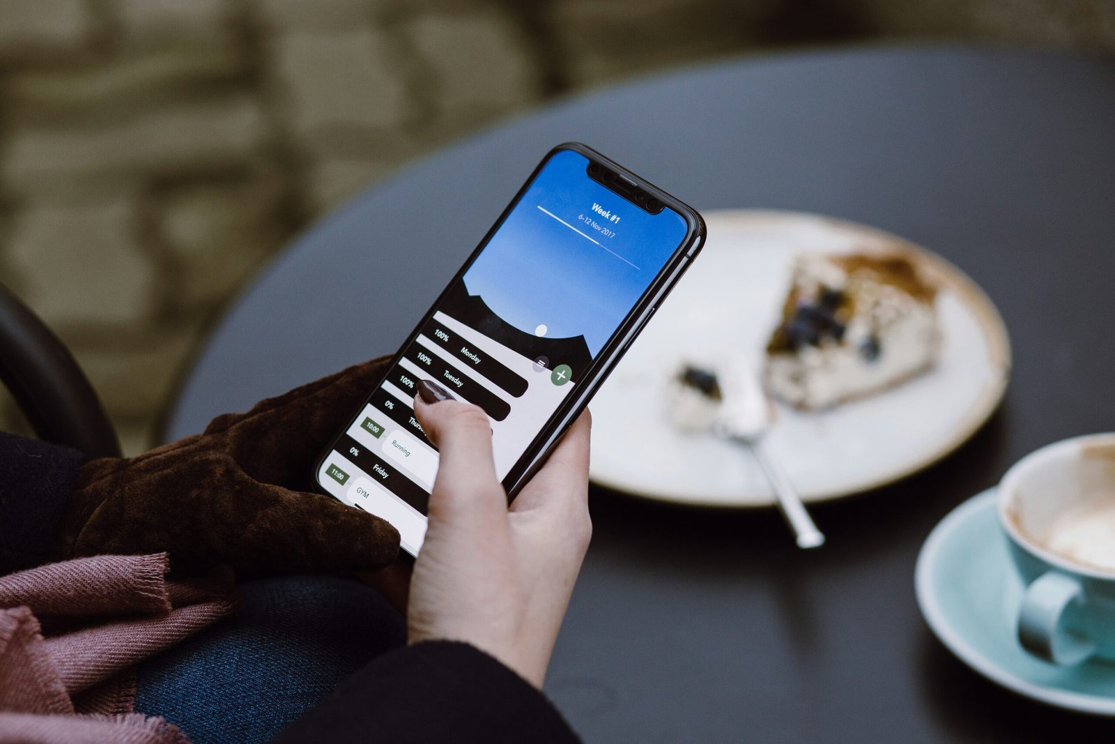 A person using a mobile phone with a productivity app open on the screen, resting on a table beside a plate with a piece of cake and a cup of coffee.