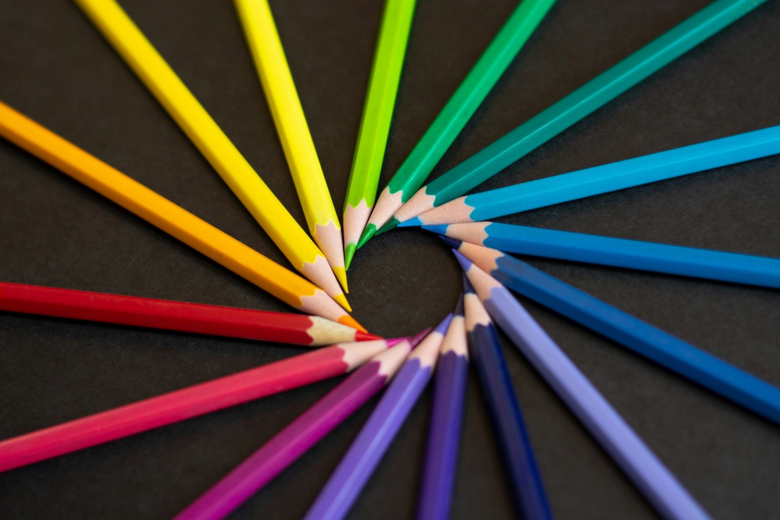 Colorful pencils arranged in a semicircle pattern on a dark background, forming a rainbow spectrum from yellow to blue to red.